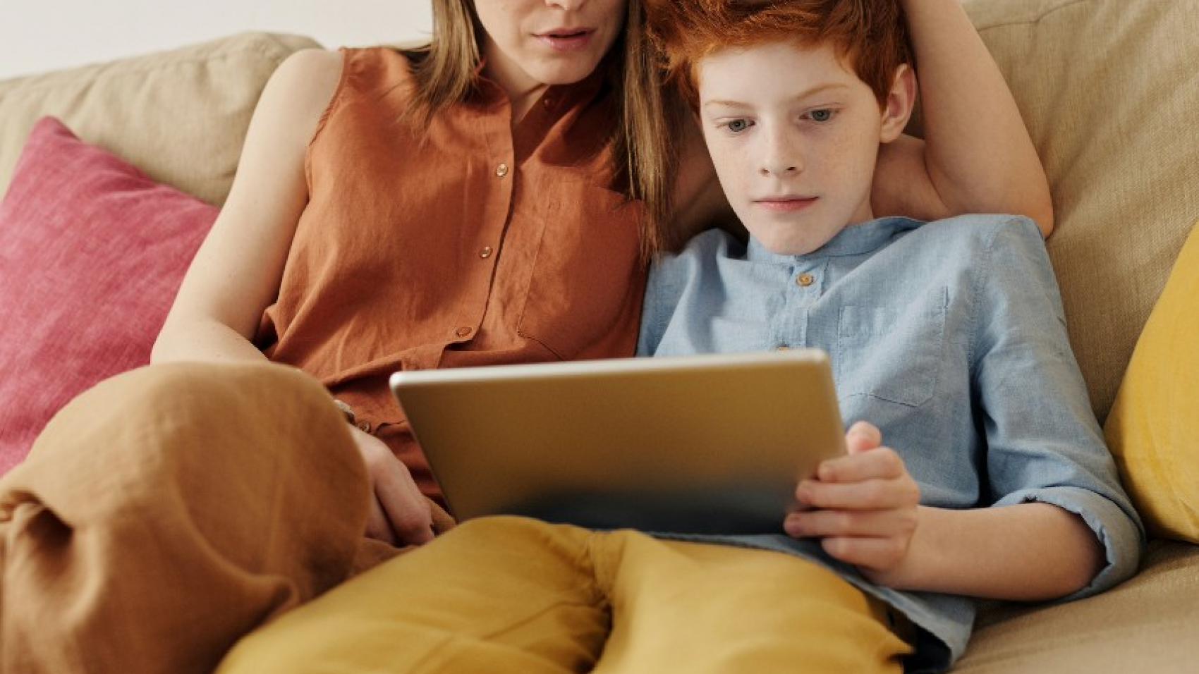 A parent sits beside her son on the couch as he looks at a tablet.