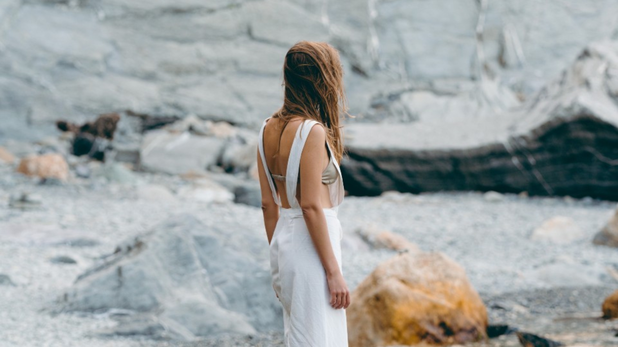 A woman stand alone on a rocky seashore, her face is not visible